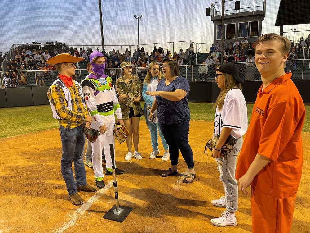 Softball and baseball players stand on the field in front of an audience. players are wearing halloween costumes including Woody and Buzz from Toy Story